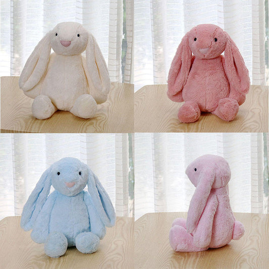 Four plush bunny toys in different colors (white, pink, blue, and pink) on a wooden surface with a light curtain background.